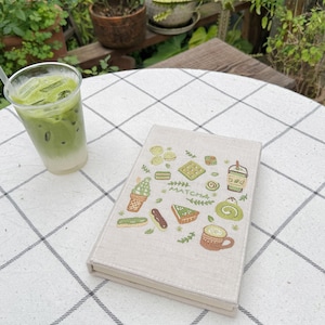 May include: A glass of iced matcha latte with a straw is next to a beige notebook decorated with matcha-themed illustrations. The notebook has drawings of matcha desserts and the word "Matcha". The items are on a white and grey checkered tablecloth.