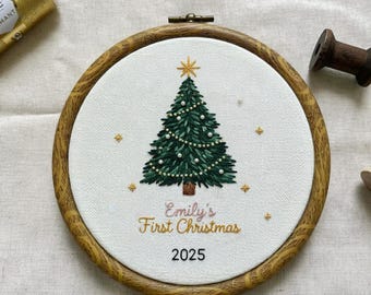 Può includere: Ricamo ad anello con albero di Natale. Il design presenta un albero di Natale verde con stella dorata e luci a catena, e il testo "Emily's First Christmas 2025" in filo d'oro. Il ricamo è in un cerchio di legno.