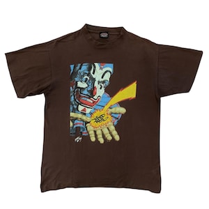 Puede incluir: Camiseta marrón con un gráfico de un payaso con pintura facial azul y roja, un rayo amarillo y texto en la mano. La camiseta es de estilo vintage, probablemente de los años 90 o principios de los 2000.