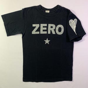 May include: Black t-shirt with the word "ZERO" in large, silver letters across the chest, and a silver star below. A white heart-shaped design is on the left sleeve. The shirt is made of a soft material.