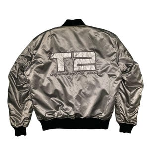 Könnte beinhalten: Silberne Satin-Bomberjacke mit schwarzem Besatz. Auf der Rückseite der Jacke steht in silbernen Blockbuchstaben der Schriftzug "T2" sowie der Text "UNIVERSAL STUDIOS JAPAN". Die Jacke hat einen gerippten Kragen, Bündchen und Saum.