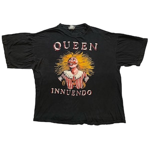 Könnte beinhalten: Schwarzes T-Shirt mit dem Wort "QUEEN" über einer Grafik einer lächelnden Figur mit wildem gelbem Haar und einem rot-weiß gestreiften Hemd. Das Wort "INNUENDO" steht darunter.