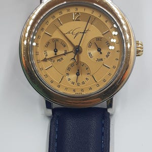 May include: A gold-toned wristwatch with a silver case and a dark blue leather band. The watch face displays multiple subdials for day, date, and month, with the brand name "Le Quat" visible. The watch has a classic, elegant design.