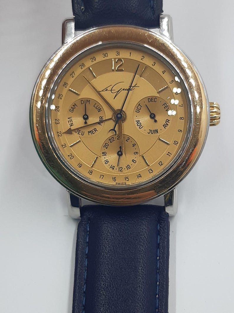 May include: A gold-toned wristwatch with a silver case and a dark blue leather band. The watch face displays multiple subdials for day, date, and month, with the brand name "Le Quat" visible. The watch has a classic, elegant design.