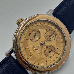 May include: A gold-toned wristwatch with a black leather band. The watch face features multiple subdials indicating the day, date, and month. The watch has a silver-colored case and a gold-colored bezel. The watch hands are black.