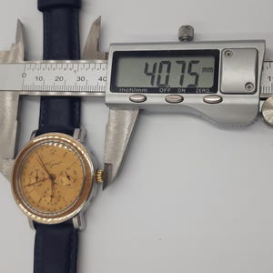 May include: A gold-toned analog wristwatch with a black leather band is measured with a digital caliper. The watch face has multiple subdials and a silver bezel. The caliper displays a measurement of 40.75 mm.