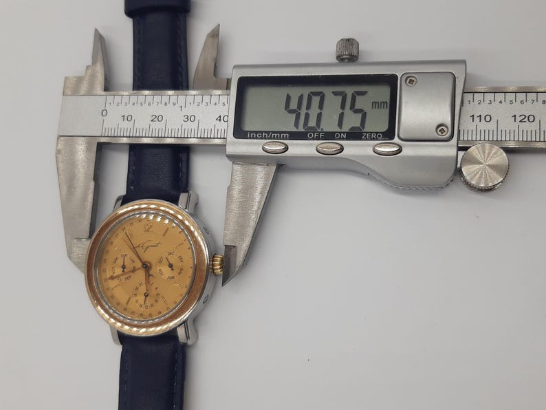 May include: A gold-toned analog wristwatch with a black leather band is measured with a digital caliper. The watch face has multiple subdials and a silver bezel. The caliper displays a measurement of 40.75 mm.