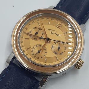 May include: A gold-toned wristwatch with a blue leather band. The watch face displays multiple subdials for day, date, and month, with Roman numerals at the hours. The watch has a stainless steel case and a textured bezel.