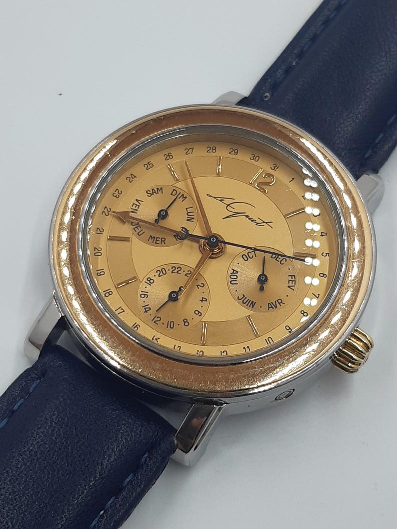 May include: A gold-toned wristwatch with a blue leather band. The watch face displays multiple subdials for day, date, and month, with Roman numerals at the hours. The watch has a stainless steel case and a textured bezel.