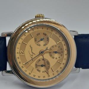 May include: A gold-toned wristwatch with a dark blue leather band. The watch face features multiple subdials for date and month, with the brand name "Le Juet" visible. The watch has a silver-colored bezel and a gold crown.