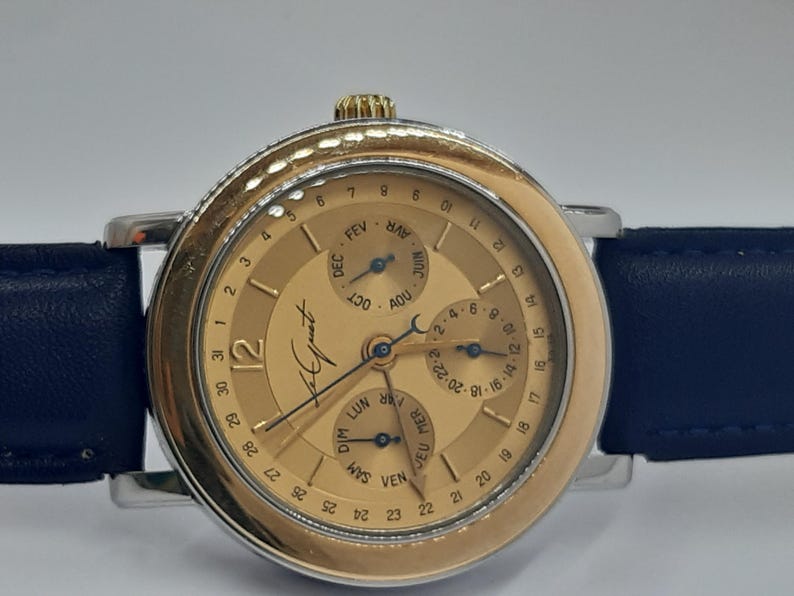 May include: A gold-toned wristwatch with a dark blue leather band. The watch face features multiple subdials for date and month, with the brand name "Le Juet" visible. The watch has a silver-colored bezel and a gold crown.