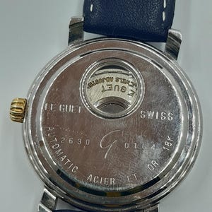 May include: A close-up of the back of a silver-toned watch with a blue leather strap. The watch face has the words "LE GUET SWISS" and "AUTOMATIC ACIER ET OR 18K" engraved. The watch also has the numbers "2630" and "0164".