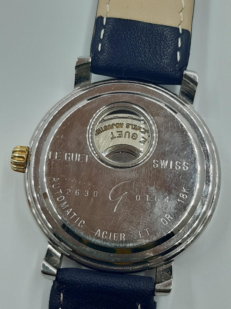 May include: A close-up of the back of a silver-toned watch with a blue leather strap. The watch face has the words "LE GUET SWISS" and "AUTOMATIC ACIER ET OR 18K" engraved. The watch also has the numbers "2630" and "0164".