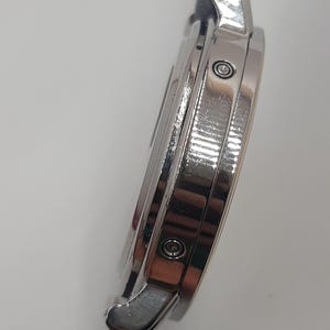 May include: A silver-toned wristwatch with a dark blue leather band. The watch case is round and features a polished finish. The side view shows the watch's intricate design, including the crown and the attachment points for the band.