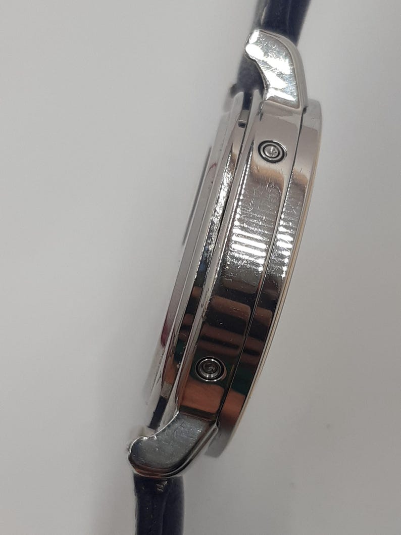 May include: A silver-toned wristwatch with a dark blue leather band. The watch case is round and features a polished finish. The side view shows the watch's intricate design, including the crown and the attachment points for the band.