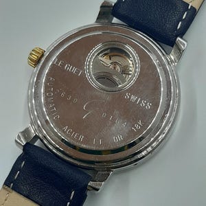May include: Close-up of the back of a silver-toned watch with a blue leather strap. The watch face is visible, displaying the inner workings and the text "LE GUET", "SWISS", and "18K OR". The strap is marked with the number "20".
