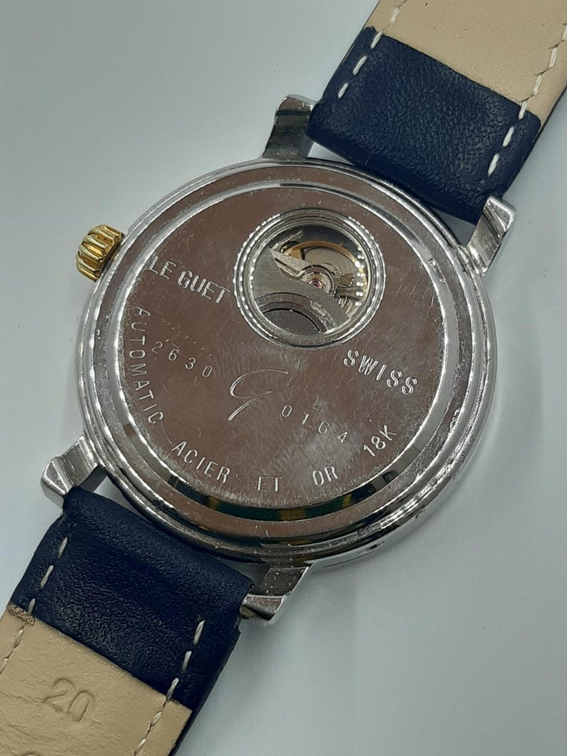 May include: Close-up of the back of a silver-toned watch with a blue leather strap. The watch face is visible, displaying the inner workings and the text "LE GUET", "SWISS", and "18K OR". The strap is marked with the number "20".
