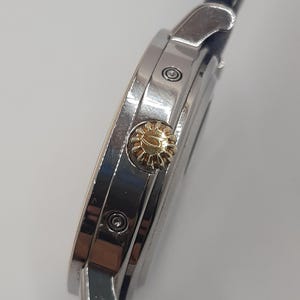 May include: Close-up of a silver wristwatch with a gold-colored crown. The watch has a dark blue leather band. The watch face is not visible. The watch is set against a white background.
