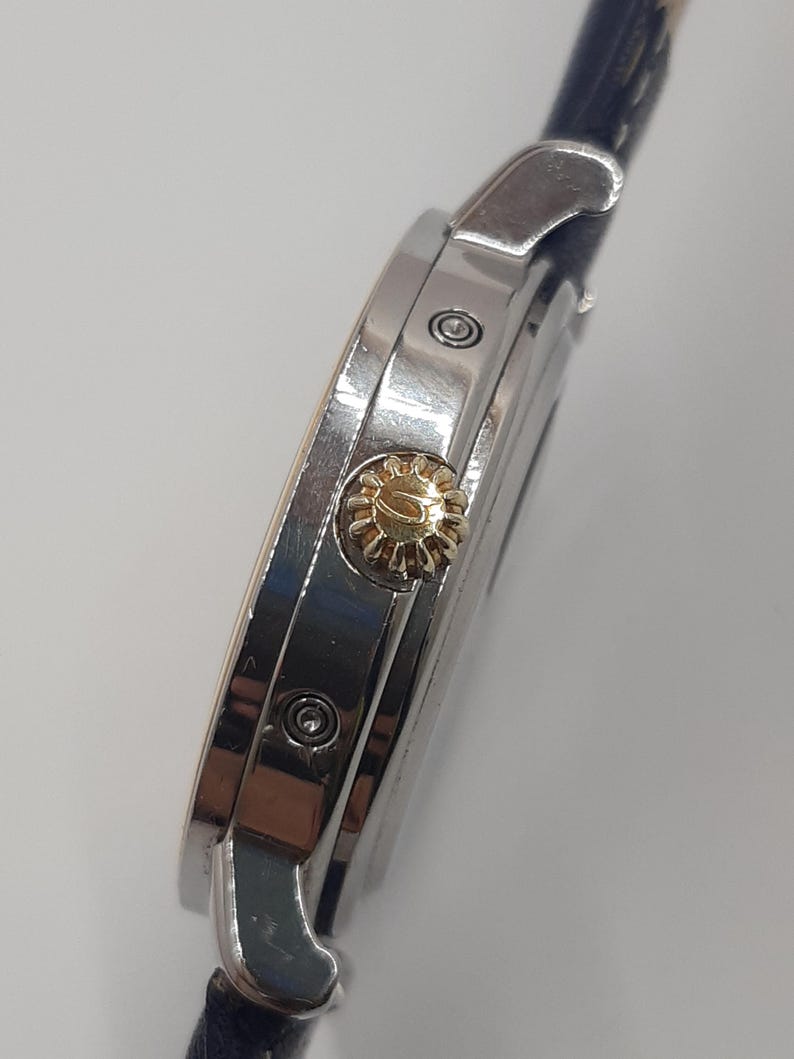 May include: Close-up of a silver wristwatch with a gold-colored crown. The watch has a dark blue leather band. The watch face is not visible. The watch is set against a white background.