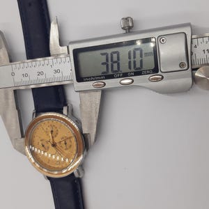 May include: A gold-toned wristwatch with a navy blue leather band is measured with a digital caliper. The watch face has multiple subdials. The caliper displays a measurement of 38.10 mm, or approximately 1.5 inches.