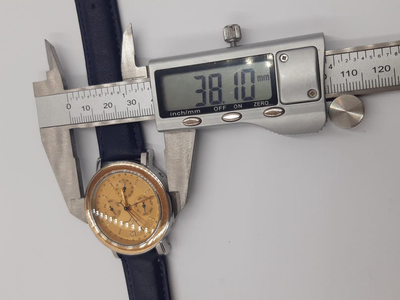 May include: A gold-toned wristwatch with a navy blue leather band is measured with a digital caliper. The watch face has multiple subdials. The caliper displays a measurement of 38.10 mm, or approximately 1.5 inches.
