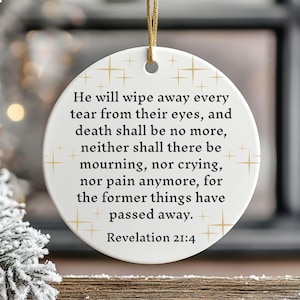 Puede incluir: Adorno navideño blanco y redondo con detalles dorados y una cuerda dorada para colgar. El adorno presenta el texto "He will wipe away every tear from their eyes...Revelation 21:4."
