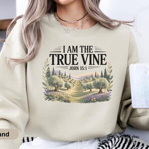 May include: Sand-colored sweatshirt with the text "I AM THE TRUE VINE JOHN 15:1" above a landscape illustration of olive trees and a path. The sweatshirt is a neutral color and has a crew neck.