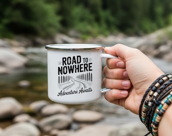 May include: White enamel camping mug with a black and white graphic that reads "Road to Nowhere, Adventure Awaits." The mug has a silver rim and handle. The mug is held by a person with multiple bracelets.