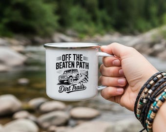 May include: White enamel camping mug with a black rim and handle. The mug features the text "OFF THE BEATEN PATH Dirt Trails" with a graphic of a 4x4 vehicle on a mountain trail. The mug is held by a person.