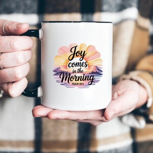 May include: White ceramic mug with a black handle and rim. The mug features a colourful floral design with the text "Joy comes in the Morning" and "Psalm 30:5". The design includes a sunrise and mountain range.