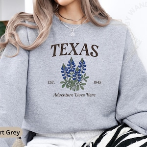 May include: A light gray sweatshirt with the word "TEXAS" in brown letters above a bluebonnet flower illustration. Below the illustration are the words "EST. 1845" and "Adventure Lives Here". The sweatshirt is sport grey.