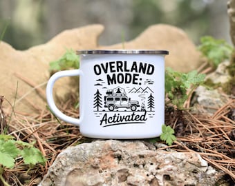 May include: White enamel camping mug with a silver rim. The mug features a black graphic with the text "OVERLAND MODE: Activated" and an illustration of a 4x4 vehicle, trees, mountains, and a campfire.