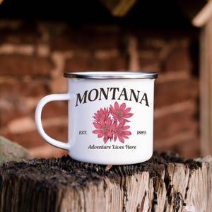May include: White enamel camping mug with a silver rim, featuring the word "MONTANA" in a serif font, with pink flowers and the text "Adventure Lives Here." The mug is set on a weathered wooden surface.
