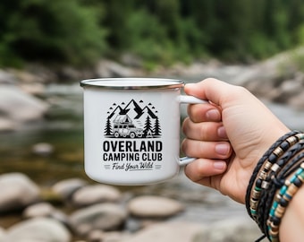May include: White enamel camping mug with a black graphic of a mountain scene, trees, and a camper van. The text on the mug reads "OVERLAND CAMPING CLUB Find Your Wild". The mug is held by a person with multiple bracelets.