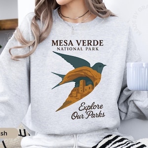 May include: Grey sweatshirt with a bird graphic incorporating Mesa Verde National Park imagery. The bird design includes the park's structures. Text reads "Mesa Verde National Park" and "Explore Our Parks".