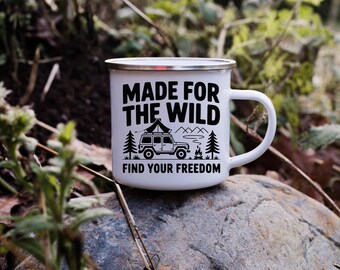 May include: White enamel camping mug with a silver rim. The mug features the text "MADE FOR THE WILD" above a graphic of a camping vehicle, trees, and mountains. Below the graphic is the text "FIND YOUR FREEDOM".