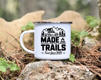May include: White enamel camping mug with a silver rim and a handle. The mug features a black graphic of a vehicle, mountains, trees, and a campfire, with the text "MADE FOR THE TRAILS" and "Find Your Wild".