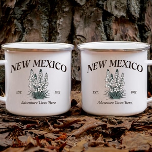 May include: Two white enamel mugs with silver rims and handles. Each mug features the text "NEW MEXICO" above a floral illustration and the words "EST. 1912 Adventure Lives Here."