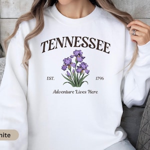 Puede incluir: Sudadera blanca con la palabra "TENNESSEE" en texto marrón arqueado. Debajo del texto hay un gráfico de flor de iris púrpura con hojas verdes. El texto "EST. 1796" y "Adventure Lives Here" también están impresos.