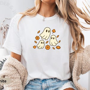 Puede incluir: Camiseta blanca con un diseño retro de tres fantasmas de dibujos animados, flores y hojas naranjas. Ideal para Halloween o el otoño. La camiseta está hecha de un material suave.