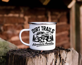 May include: White enamel mug with a black and white graphic. The mug features the words "Dirt Trails Adventure Awaits" with an illustration of a 4x4 vehicle, trees, and mountains. The mug has a silver rim and a white handle.