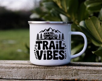 May include: White enamel camping mug with a black design featuring a mountain range, trees, and a 4x4 vehicle. The words "TRAIL VIBES" are printed in a bold, retro font, with floral accents.
