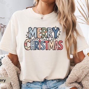 以下が含まれることがあります： 「MERRY CHRISTMAS」の文字が装飾的なフォントでプリントされたアイボリー色のTシャツ。テキストは花柄とストリングライトで飾られています。ホリデーをテーマにしたアパレルコレクションの一部です。