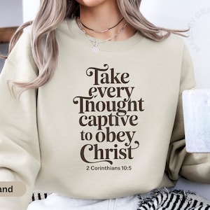 May include: Sand-colored sweatshirt with the text "Take every thought captive to obey Christ" in a dark brown script. The sweatshirt has a crew neck and long sleeves. The text is from 2 Corinthians 10:5.