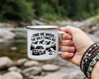 May include: White enamel camping mug with a black graphic of a mountain range, trees, a 4x4 truck, and a tent. The text on the mug reads "FIND ME WHERE THE WILD THINGS ARE". The mug has a silver rim and handle.