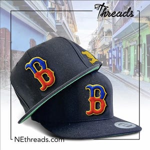 May include: Two navy blue baseball caps with a green brim. Each cap features a large, embroidered "B" logo in red, yellow, and blue. The text "NEthreads.com" is visible on the bottom left corner. The top right corner has the text "NE Threads".