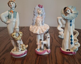 May include: Three porcelain figurines standing in a row. The left figure wears a striped hat and holds a large gold key. The center figure has long hair, a patterned dress, and a small dog. The right figure wears a blue ruffled collar and robe.