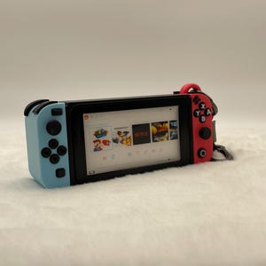May include: A handheld gaming console with a black screen displaying game icons. The console has a light blue left side with a joystick and buttons, and a red right side with buttons. A black frame surrounds the screen.
