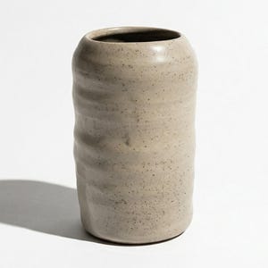 May include: A cylindrical ceramic vase in a neutral beige color with a textured, ridged surface. The vase has a dark interior and is set against a white background, casting a shadow.