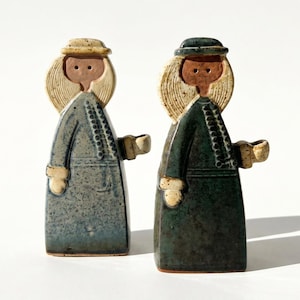 May include: Two ceramic figurines of figures in long coats and hats. One is blue-grey, the other is dark green. Both have blonde hair and brown faces. The figures have small, curved arms and are standing on a white surface.